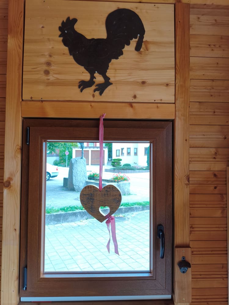 Fenster der Hütte mit Hahn-Silhouette darüber und einem Holzherz
