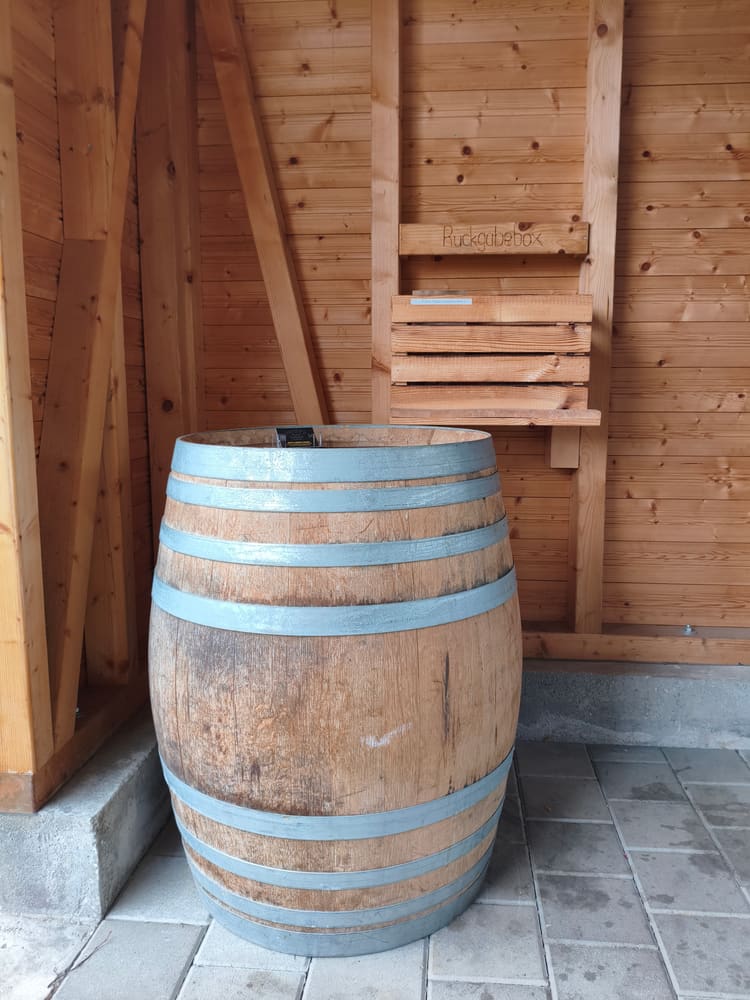 Holzfach mit der Aufschrift „Rückgabebox“ an der Hüttenwand, davor ein Weinfass